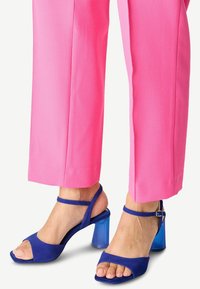 Blå mocka högklackade sandaler med en genomskinlig blå blockklack och ankelrem, tillsammans med livliga rosa vida byxor.