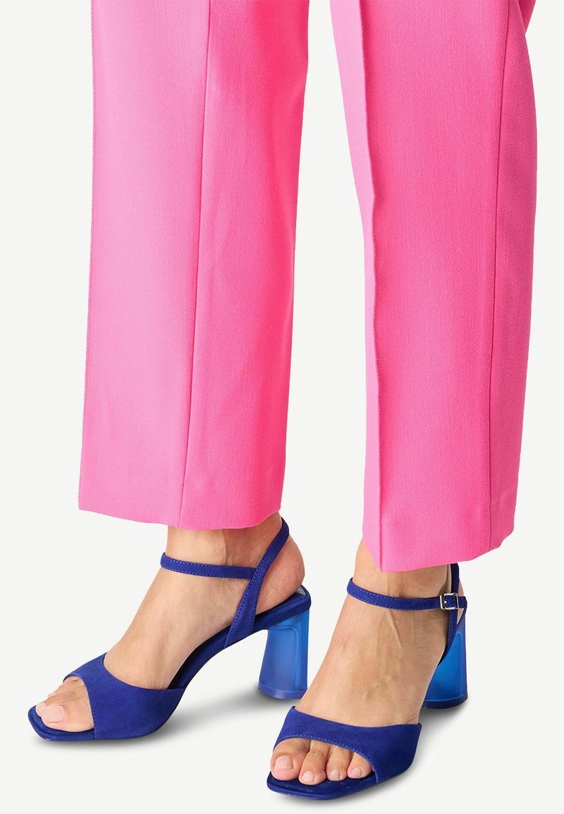 Blå mocka högklackade sandaler med en genomskinlig blå blockklack och ankelrem, tillsammans med livliga rosa vida byxor.