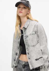 Jasnoszara, dżinsowa kurtka o skróconym fasonie, z kieszenią z przodu i czarnym akcentem logo. Nosi ją na dopasowanej, czarnej bluzce. Szara czapka dopełnia stylizację.