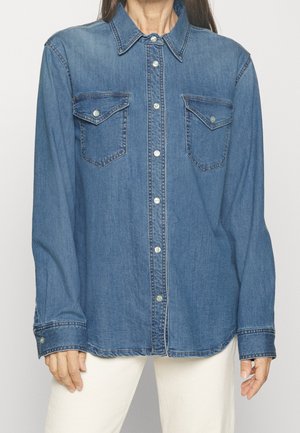 Vrouw draagt een blauwe denim blouse met knopen en twee borstzakken, en witte broek, staand tegen een effen achtergrond.