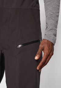 Juodas audinys, turintis sklandžią tekstūrą, su šonine užtrauktukine kišenė ir kontrastiniu pilku ilgomis rankovėmis viršumi. Artimas rankos planas.