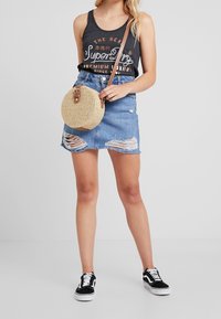 Borsa a tracolla rotonda con una tracolla marrone, abbinata a una canotta grigia con logo e a una mini gonna di jeans con dettagli strappati. Sneakers nere completano il look.