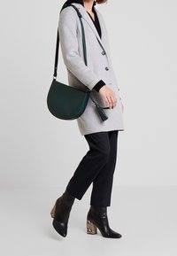 Borsa crossbody in pelle verde a forma semicircolare, caratterizzata da un dettaglio con frange, indossata con un cappotto grigio chiaro e pantaloni neri.
