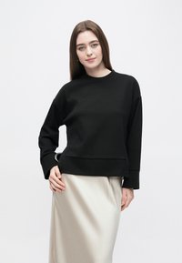 Μαύρο φούτερ με μακριά μανίκια και στρογγυλή λαιμόκοψη. Μαλακό ύφασμα, άνετη εφαρμογή, με τσέπη και διακοσμητικό μεταλλικό στοιχείο.