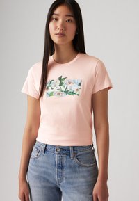 T-shirt en coton rose clair à manches courtes, avec un motif floral en vert et bleu, centré avec un logo blanc "Levi's".