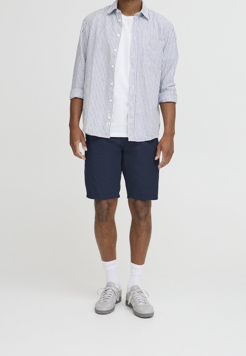 Homme portant un t-shirt blanc, une chemise à rayures bleues et blanches boutonnée, un short bleu marine, des chaussettes blanches et des baskets grises, debout devant un fond blanc.