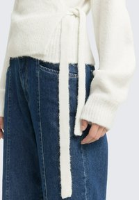 Bela pletena jopica z detajlom z vezanjem v pasu, v kombinaciji z modrimi jeans hlačami. Jopica ima rebraste manšete in mehak občutek.