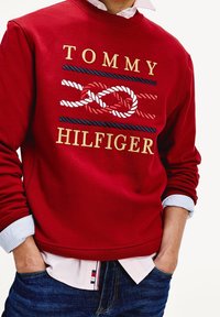 Red sweatshirt with "TOMMY HILFIGER" embroidered in gold and blue, featuring a rope design. Worn over a light blue collared shirt.
