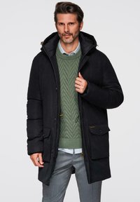 Černá hřejivá parka s kapucí, se zapínáním na zip, předními kapsami a žlutými tkaničkami. Nositela ji má přes zelený pletený svetr.