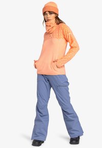 Oranje fleece top met patroonmouwen en een hoge kraag, gecombineerd met lichte blauwe broek en zwarte laarzen. Gehaakte oranje beanie inbegrepen.