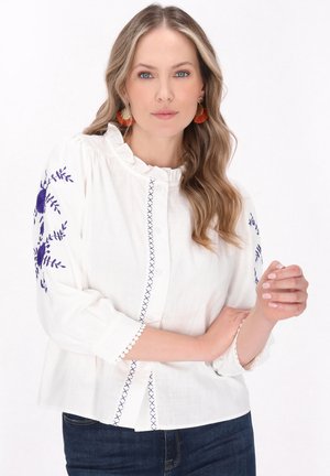 Camicia bianca con collo a balze e ricamo floreale blu sulle maniche. Presenta pizzo sui polsini e una vestibilità comoda.