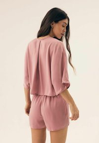 Różowy satynowy kombinezon z krótkimi rękawami, luźnym krojem i detalem w postaci pliski z tyłu, prezentujący miękką teksturę i delikatny połysk.