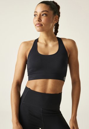 Frau mit geflochtenem Haar, die einen schwarzen Sport-BH und hoch taillierte schwarze Leggings trägt und nach links schaut vor hellem Hintergrund.
