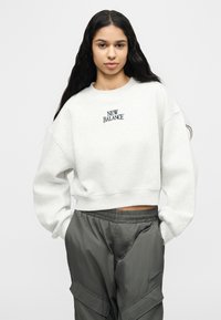 Γυναίκα με μακριά σκούρα μαλλιά φορώντας ανοιχτό γκρι cropped φούτερ New Balance και σκουρό γκρι παντελόνι cargo με τα χέρια στις τσέπες.