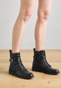 Bottes à cheville en cuir noir avec un design matelassé, des clous dorés et une accent de boucle. Bout carré et talon bas avec un système de laçage et une fermeture éclair.