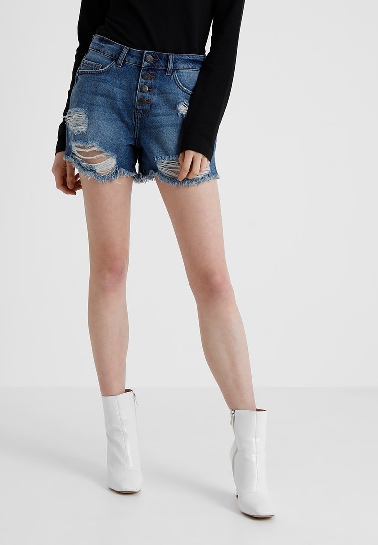 Osoba v potrhaných modrých džínech, černém dlouhém rukávu a bílých kotníkových botách stojí před jednobarevným pozadím.