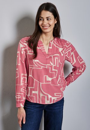 Rosa Bluse mit geometrischen cremefarbenen Mustern, ausgestattet mit einem V-Ausschnitt und langen, gerafften Ärmeln. Kombiniert mit dunklen Jeans.