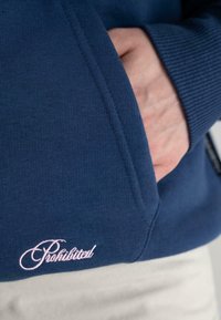 Felpa blu navy con texture a costine, tasca frontale e ricamo "Proibito" in rosa all'orlo.