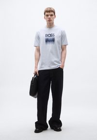 Valgehall lühikeste varrukatega T-särk, millel on "BOSS" logo ja laineline graafika, kombineeritud tumedate laiade pükstega, hoides musta kotti, neutraalsel taustainternet.