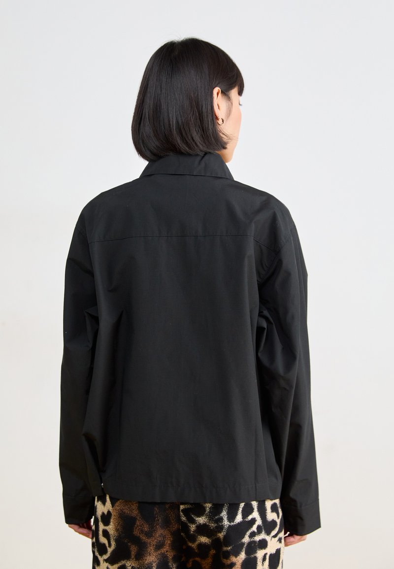 Veste noire avec un col, manches longues et coupe décontractée. Texture lisse, détail de couture dans le dos, portée sur un pantalon à imprimé léopard.