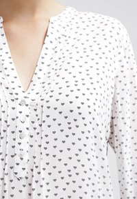 V-paita, jossa on valkoinen väri ja pienen mustan raitasydämen kuvio, neljä nappia ja laskosteet edessä. Kevyt kangas.