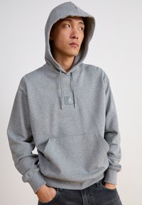 Sudadera gris de material suave, con un bolsillo frontal, cordón ajustable y parche con logo en el pecho. Diseño con capucha.