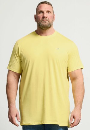 Muskuløs mand med skæg iført simpel gul kortærmet t-shirt og blå jeans, stående mod en ensfarvet lys baggrund.