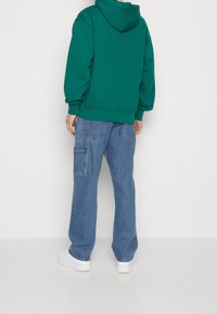 Grüne Kapuzenjacke, blaue weite Jeans mit Cargo-Taschen und weiße Sneakers. Weicher Stoff, lockerer Schnitt, lässiges Design.