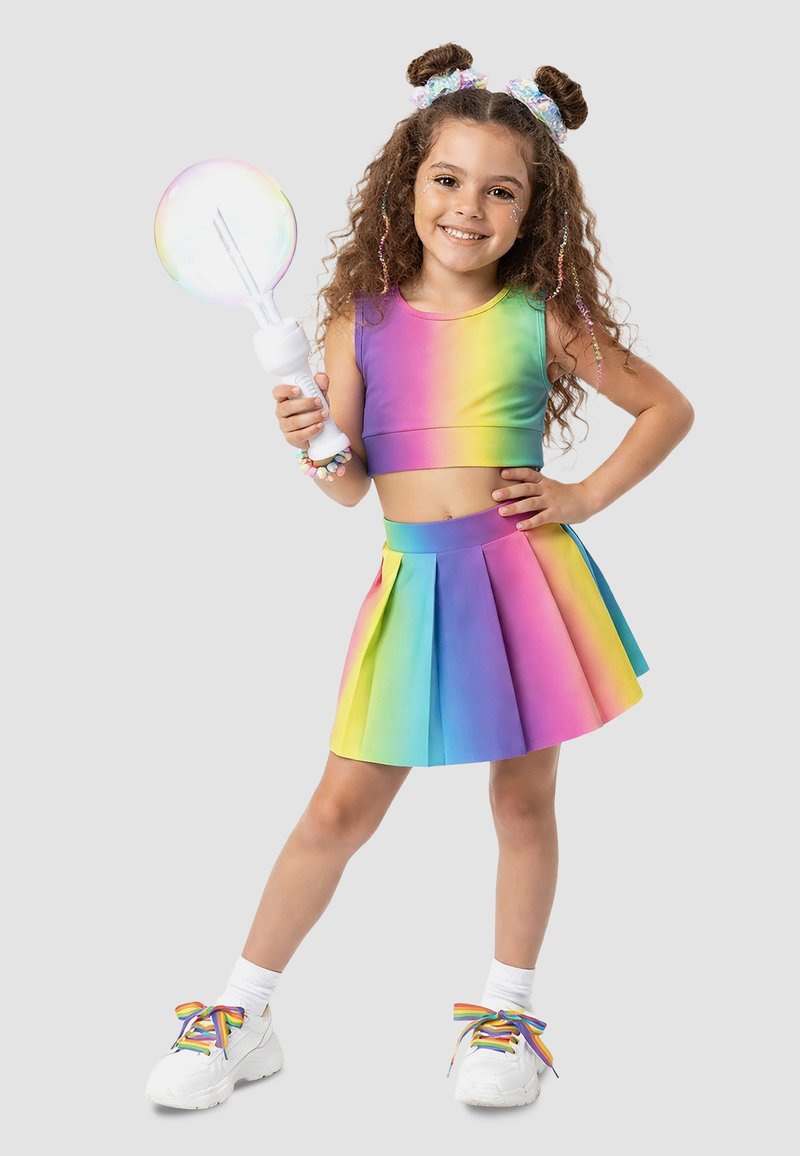 Ung pige smiler, iført regnbue crop top og nederdel, holder en bobleblæser med iriserende boble, regnbue snørebånd på hvide sneakers.