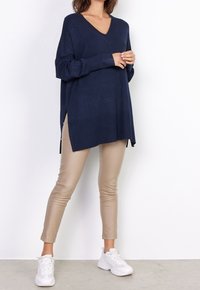 Maritimer V-Ausschnitt-Pullover mit langen Ärmeln, seitlichen Schlitzen. Kombiniert mit beigefarbenen, lederähnlichen, eng anliegenden Hosen und weißen Turnschuhen. Glatte Textur.