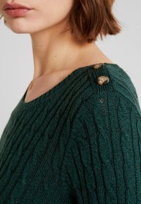 Suéter verde de punto con patrón trenzado, escote ancho y dos botones decorativos en el hombro. La textura parece suave y acogedora.