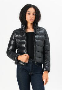 Zwarte pufferjack met een hoge kraag, glanzende afwerking, ritssluiting en horizontaal gestikte ontwerp. Draagt over een zwart top en jeans.
