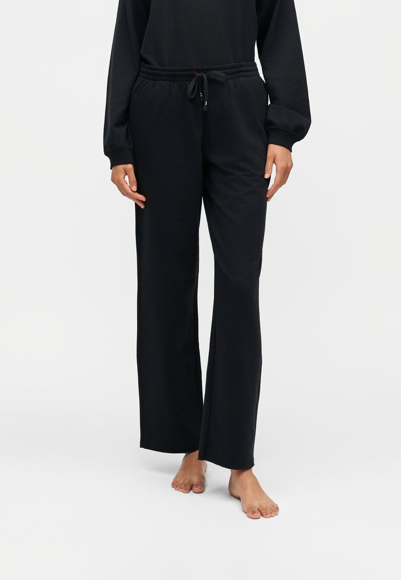 Personne portant un pantalon noir à jambes larges et un haut noir à manches longues assorti, debout pieds nus sur un fond blanc.