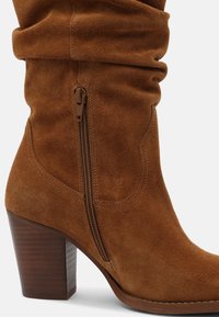 Brązowy zamszowy kozaki sięgające do kolan z marszczonym wykończeniem na górze, bocznym zamkiem błyskawicznym i masywnym drewnianym obcasem, z przeszyciami podkreślającymi kontury.