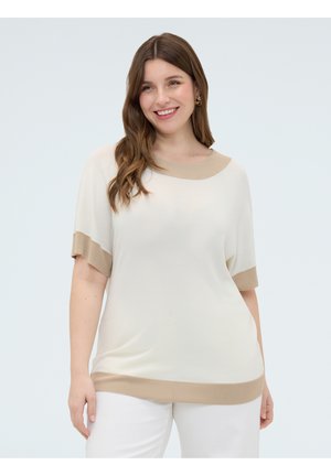 Lächelnde Frau mit langen braunen Haaren, die ein lockeres cremefarbenes Oberteil mit beigem Besatz und weiße Hosen trägt, steht vor einem einfarbigen Hintergrund.