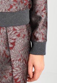 Bordeaux kledingstuk met een bloemenpatroon in zilver, voorzien van een grijze geribbelde manchette. Close-up van de arm en mouwdetails.