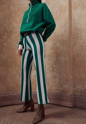 Persoon draagt een groene trui met halve rits, groene en witte vertically gestreepte hoge taille broek, en bruine enkellaarsjes met hak, staand binnen.