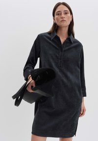 Vrouw in een donker corduroy shirtjurk die een zwarte gestructureerde handtas vasthoudt, staand tegen een effen lichte achtergrond.