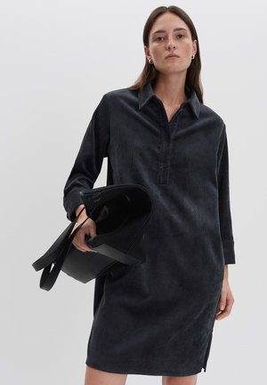 Donna in un vestito camicia in corda scura che tiene una borsa nera testurizzata, in piedi contro uno sfondo chiaro e uniforme.