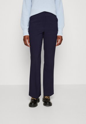 Personne portant un pantalon droit bleu marine, un pull en tricot bleu clair et des mocassins noirs avec détail chaîne dorée, debout devant un fond uni.