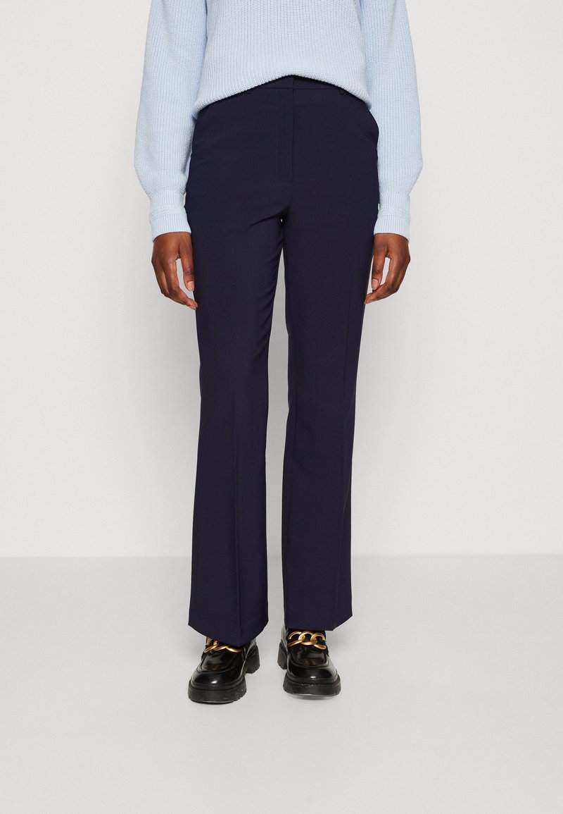 Personne portant un pantalon droit bleu marine, un pull en tricot bleu clair et des mocassins noirs avec détail chaîne dorée, debout devant un fond uni.
