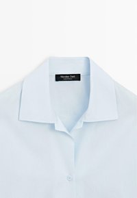 Lichtblauw overhemd met kraag, voorzien van een enkele zichtbare knop en een zwart etiket met de tekst "Massimo Dutti Made in Portugal" aan de binnenkant van de kraag.