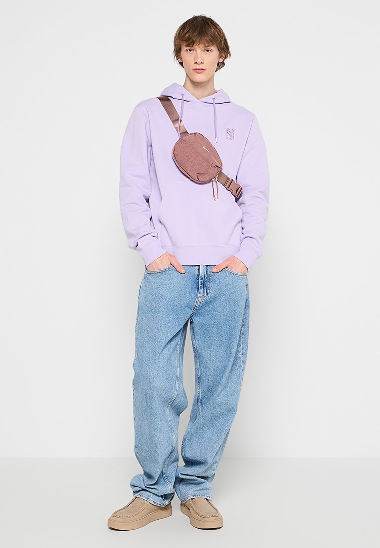 Hellblaues Hoodie, lässige blaue Jeans und beigefarbene Slipper. Braune Umhängetasche mit abgerundeter Form und Reißverschluss.