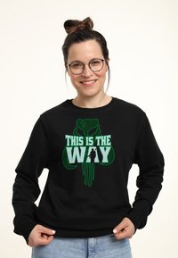 Sudadera negra con diseño gráfico verde que presenta el texto "ESTE ES EL CAMINO" y una silueta. Textura suave, cuello redondo, ajuste clásico.