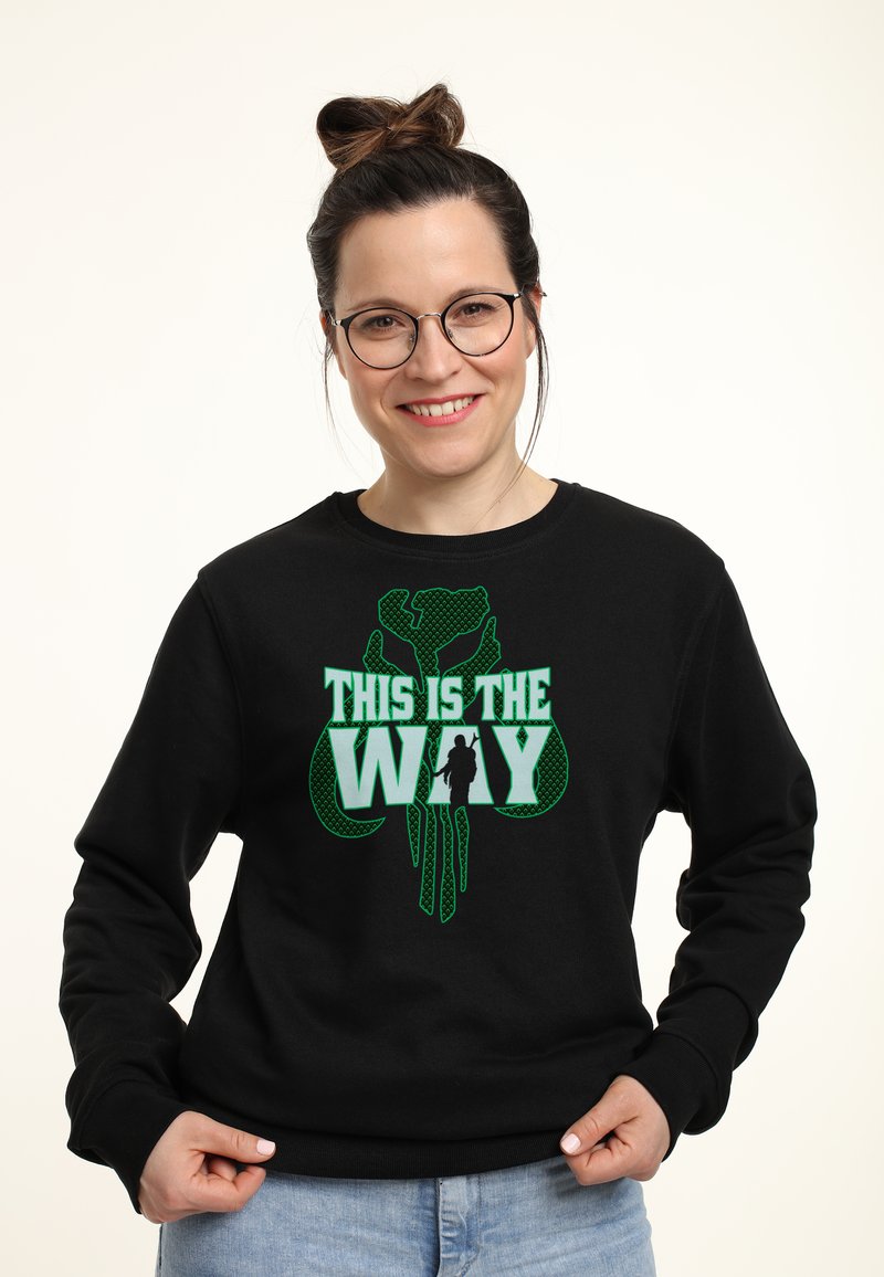 Sudadera negra con diseño gráfico verde que presenta el texto "ESTE ES EL CAMINO" y una silueta. Textura suave, cuello redondo, ajuste clásico.
