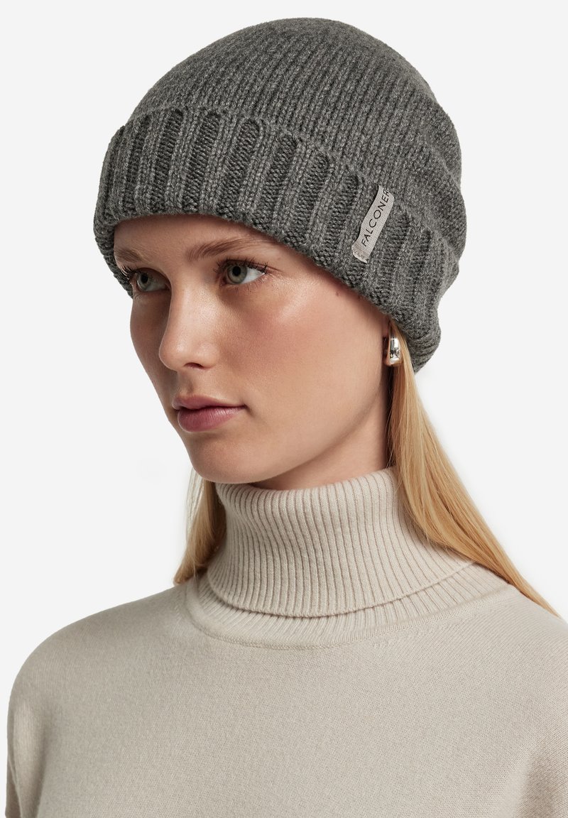 Donna dalla pelle chiara e capelli biondi indossa un berretto lavorato a maglia grigio con l'etichetta Falconeri e un maglione a collo alto beige a coste.