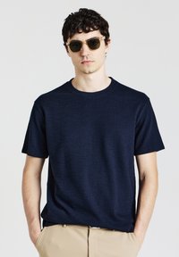 T-shirt a girocollo in tessuto testurizzato di colore navy, realizzato in un tessuto leggero, abbinato a occhiali da sole trasparenti e pantaloni beige.