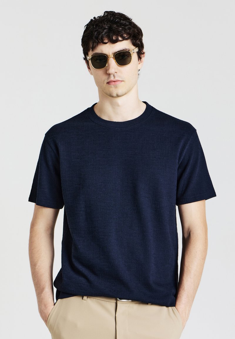 T-shirt a girocollo in tessuto testurizzato di colore navy, realizzato in un tessuto leggero, abbinato a occhiali da sole trasparenti e pantaloni beige.