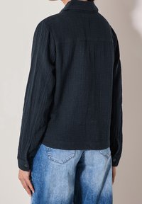 Chemise à manches longues en tissu texturé bleu marine foncé, avec un col à revers et des poignets boutonnés, associée à un jean bleu clair.