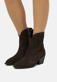 Stivali alla caviglia in suede marrone scuro con tacco basso impilato e cerniere laterali indossati su gambe nude contro uno sfondo semplice.